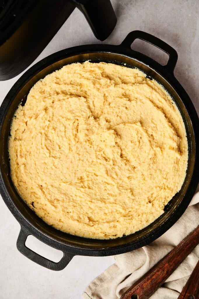 A round baking dish filled with uncooked air fryer cornbread batter sits on a light countertop, with a wooden spoon resting nearby.
