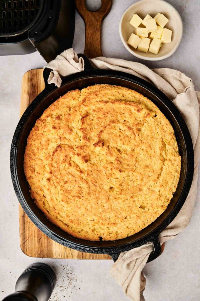 A freshly baked air fryer cornbread, golden and warm, sits in a cast iron skillet on a wooden board, accompanied by creamy butter cubes.