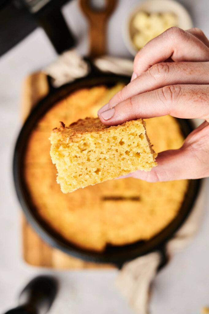 A hand holds a piece of cornbread, perfectly golden and crumbly, with a cast iron skillet brimming with air fryer cornbread in the background.