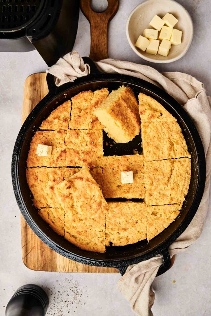 An air fryer cornbread, perfectly sliced and topped with butter squares, rests in a cast iron skillet on a wooden board. Nearby, a small bowl brims with additional butter cubes.