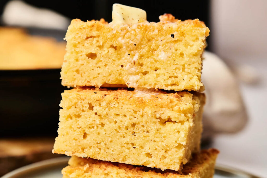 A stack of air fryer cornbread slices sits on a plate, each piece perfectly golden with a pat of butter melting enticingly on top.