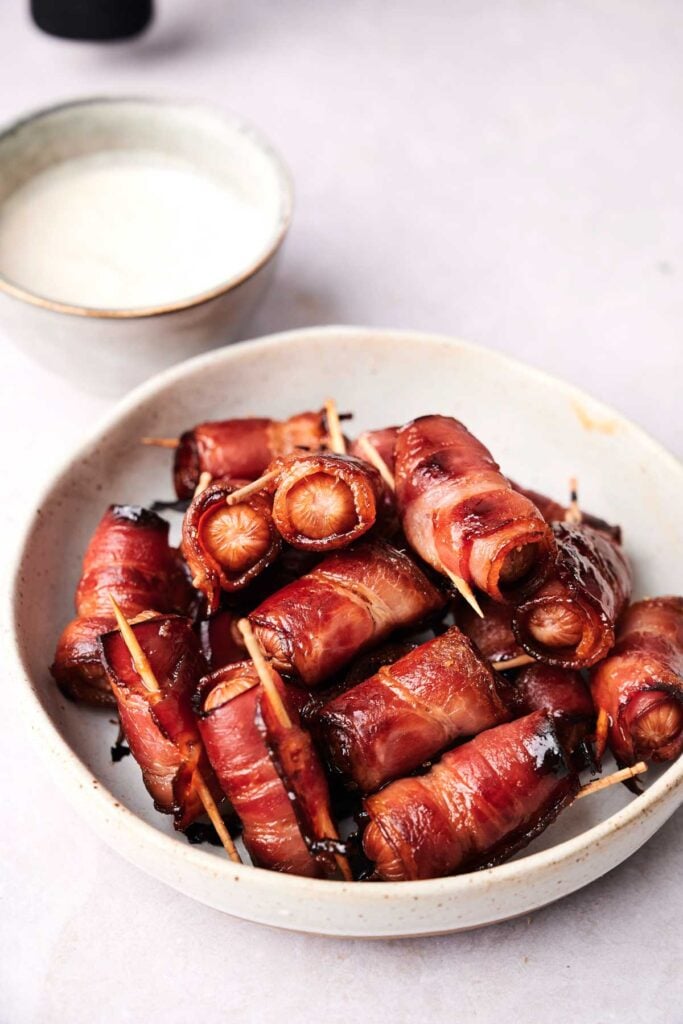 A bowl of bacon-wrapped sausages held with toothpicks, next to a bowl of dipping sauce on a light surface.