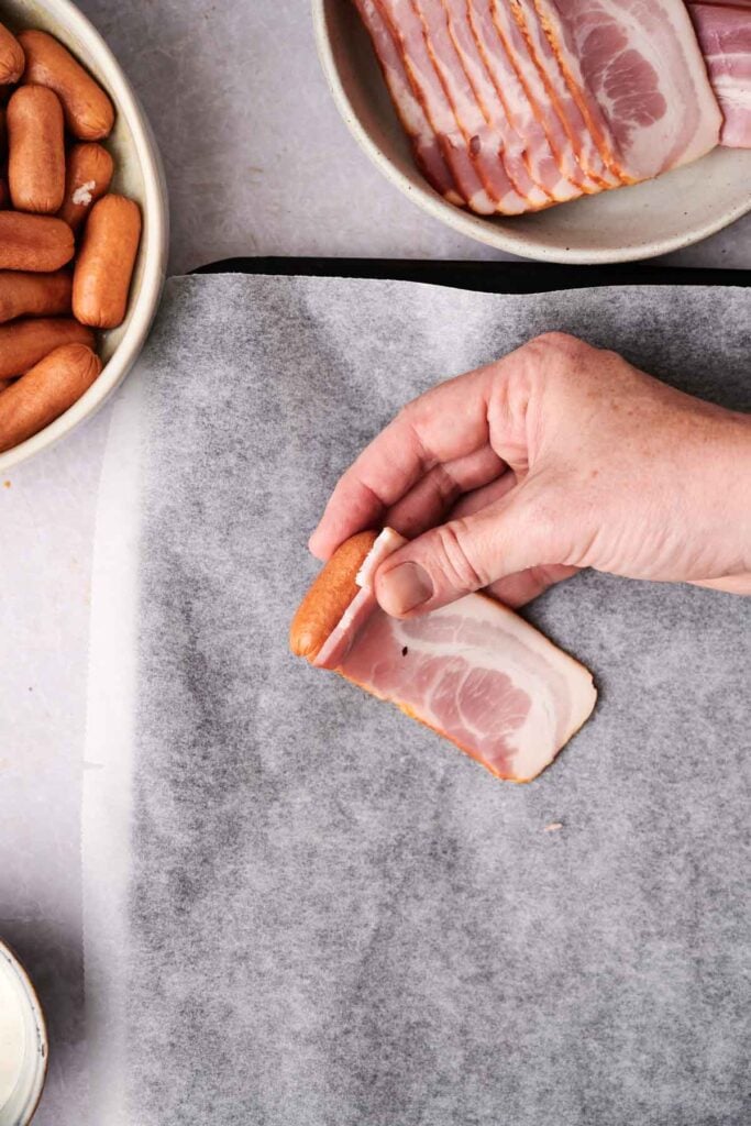 A hand wrapping a small sausage in a slice of bacon on a baking sheet. Surrounding the hand are bowls containing more sausages and bacon.