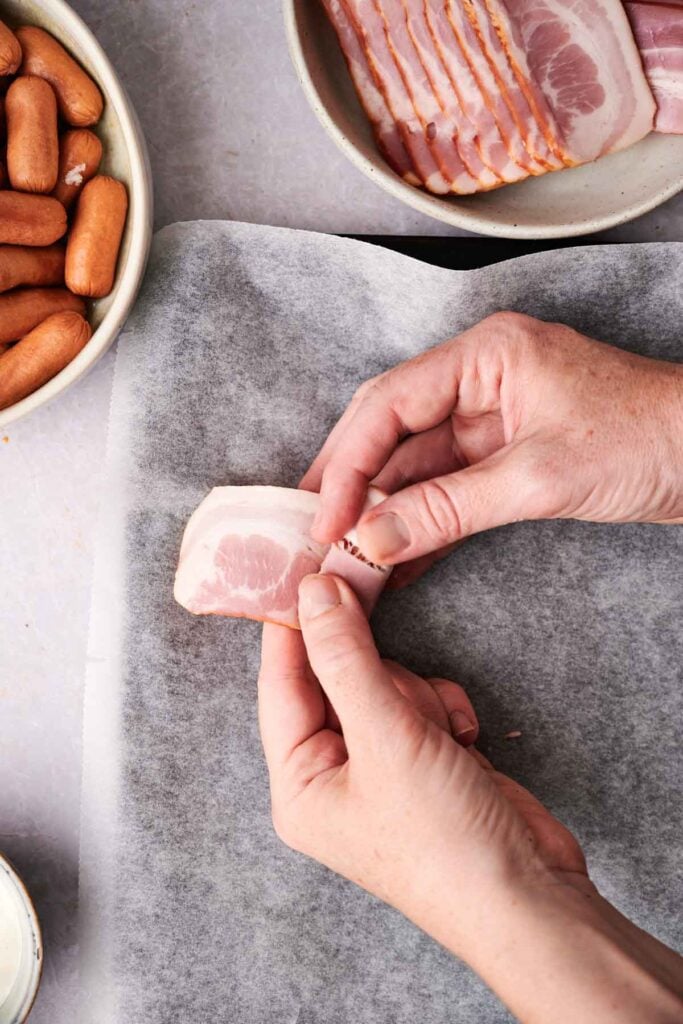 Hands rolling a slice of bacon on wax paper with a bowl of mini sausages and more bacon slices nearby.