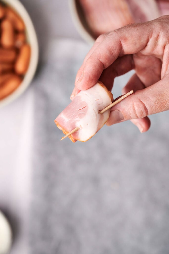 A hand holding a piece of rolled deli meat with a toothpick, with a blurred background of a bowl and cloth.