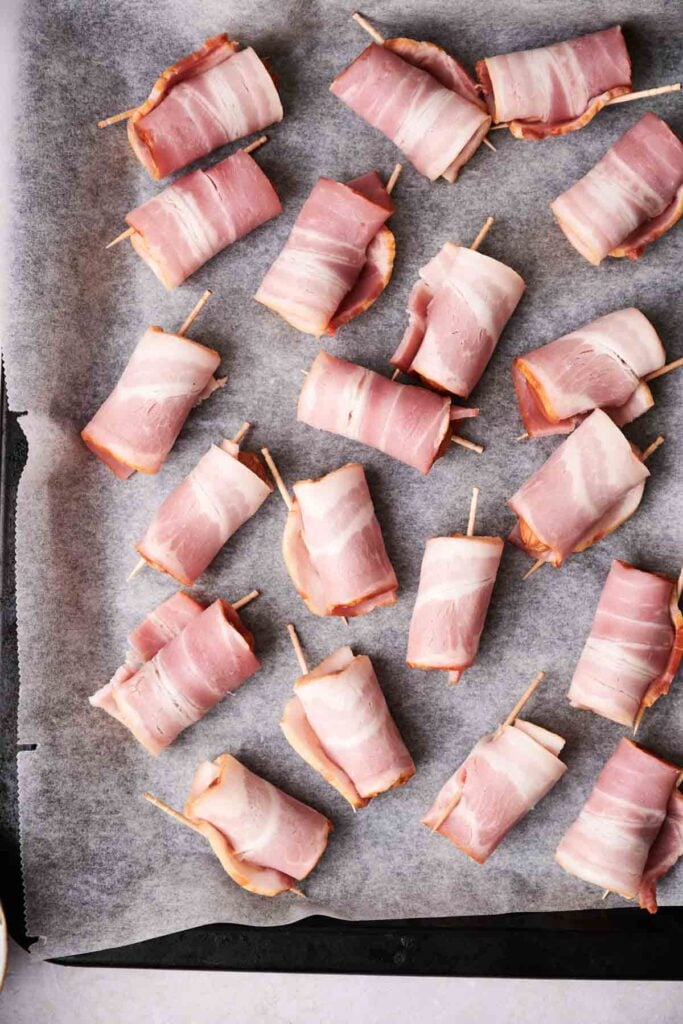 Bacon-wrapped bites secured with toothpicks are arranged on a parchment-lined baking sheet.
