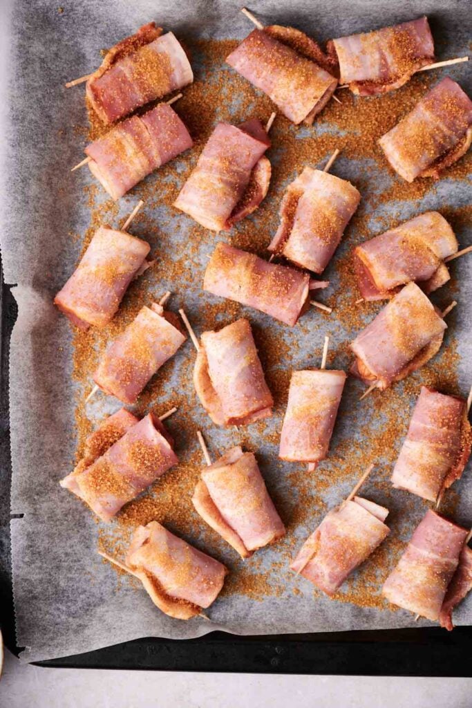 Bacon-wrapped snacks on a baking sheet with parchment paper, secured with toothpicks, sprinkled with seasoning.