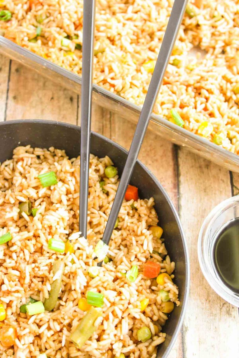 A bowl of oven-baked fried rice with vegetables sits invitingly next to a glass dish brimming with more flavorful rice, complemented by a small bowl of soy sauce and chopsticks for the perfect dining experience.