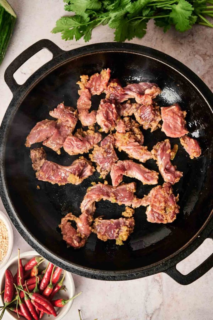 Sliced beef cooking in a black pan next to a bowl of red chilies and fresh herbs on a light countertop.