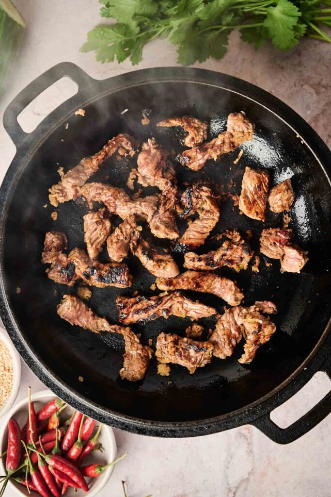 Strips of grilled meat in a black pan on a marble countertop, with a bowl of red chili peppers and fresh herbs nearby.