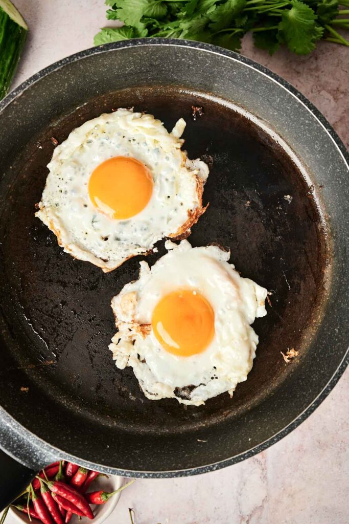 Two sunny-side-up eggs cooking in a black frying pan on a countertop, with cilantro and red chilies nearby.