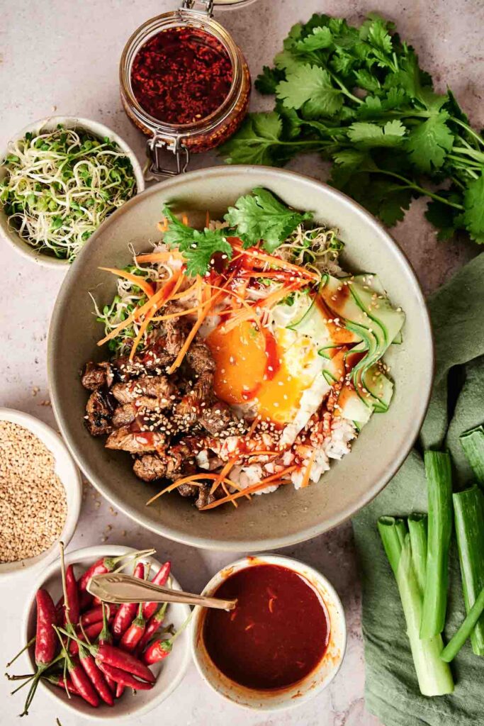 A bowl with seasoned beef, shredded carrots, cucumber, avocado, sprouts, and a soft-boiled egg, surrounded by fresh chili, sauces, sesame seeds, and cilantro leaves.