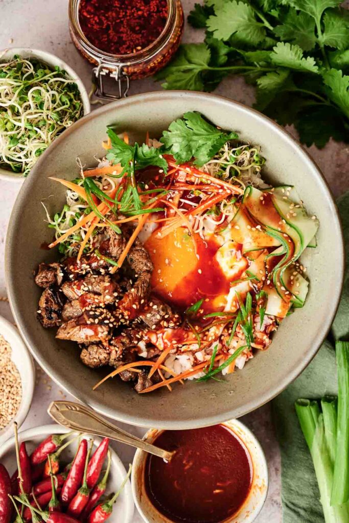A bowl of bibimbap with beef, vegetables, and egg, topped with sesame seeds, surrounded by ingredients like chili, sprouts, and sauces.