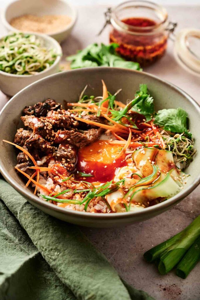 Bowl of bibimbap with rice, beef, vegetables, egg yolk, and sauce. Toppings include carrots, sprouts, cucumber, and cilantro. Side dishes and condiments visible in the background.