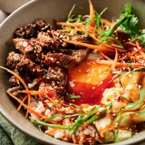 A bowl of rice topped with sliced beef, a runny egg, carrots, cucumber, greens, and sesame seeds, drizzled with a red sauce.