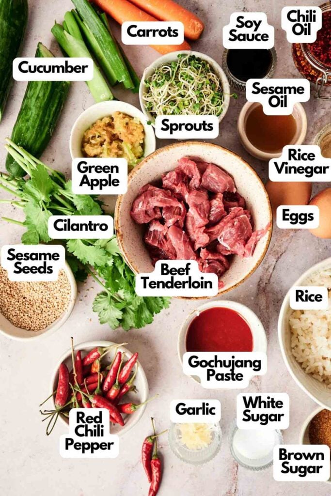 Ingredients for a Korean dish are arranged on a table, including beef tenderloin, rice, garlic, soy sauce, chili oil, sesame oil, green apple, cilantro, sprouts, carrots, cucumbers, and spices.