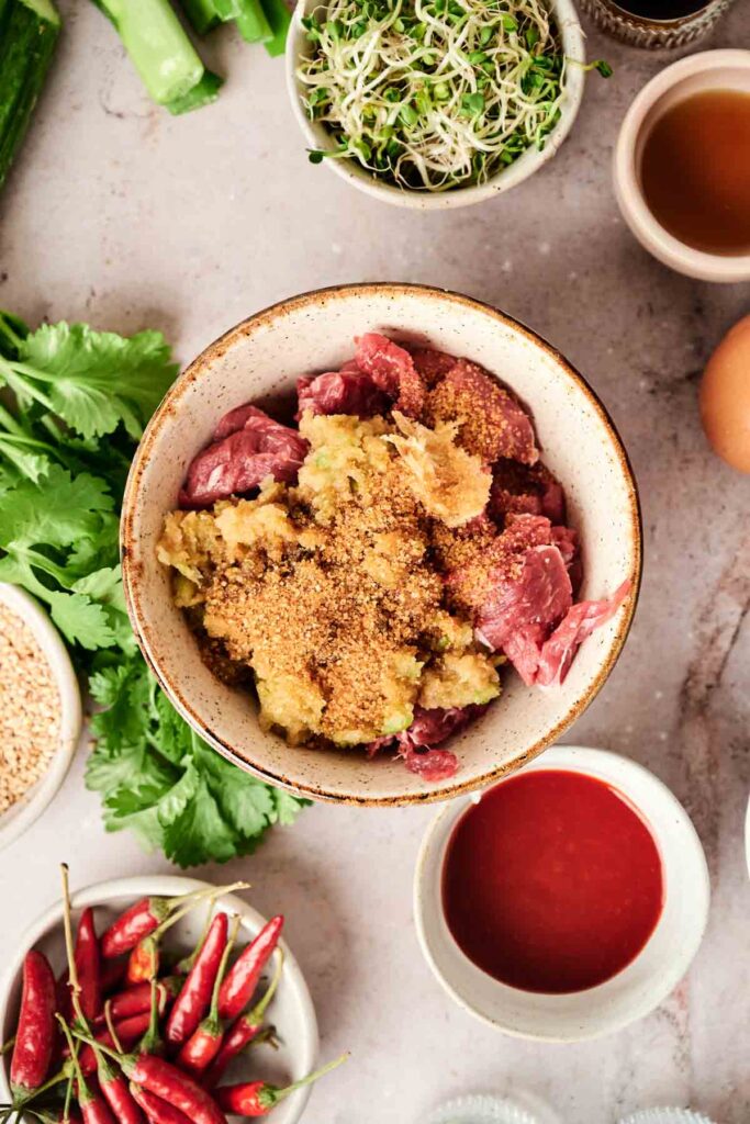 A bowl with meat and mashed ingredients surrounded by chili peppers, herbs, sprouts, a whole egg, and bowls of spices and sauces on a stone surface.