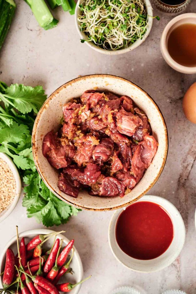 A bowl of marinated beef surrounded by fresh cilantro, red chilies, sprouts, an egg, sesame seeds, a sauce, and green onions on a countertop.