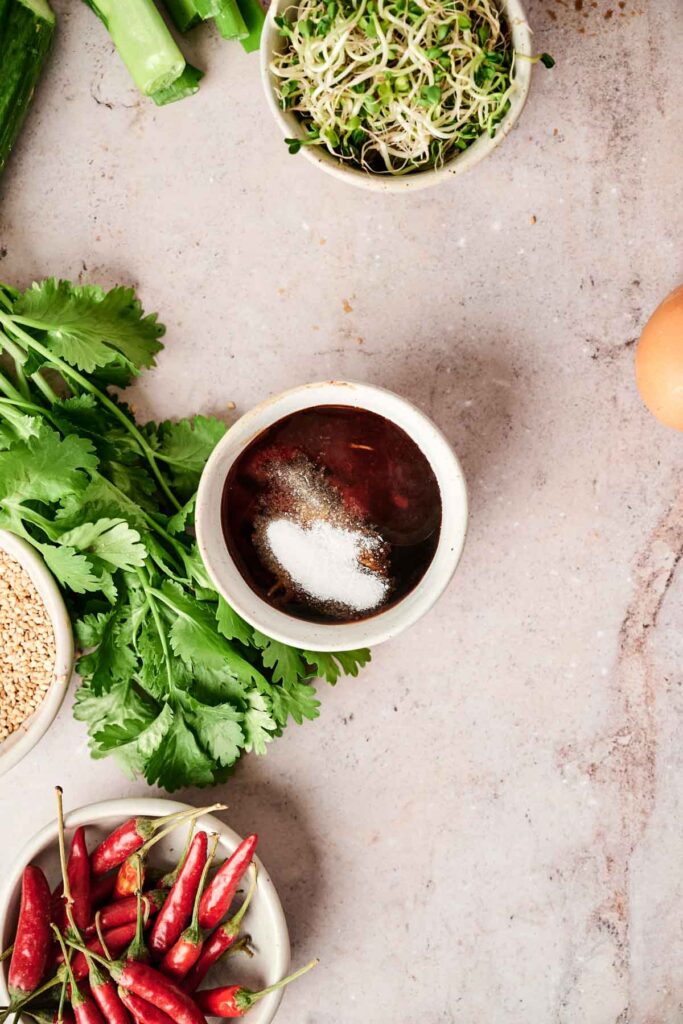 A bowl of sauce with sugar on top, surrounded by cilantro, red chili peppers, sprouts, and a partial view of an egg on a light-colored surface.