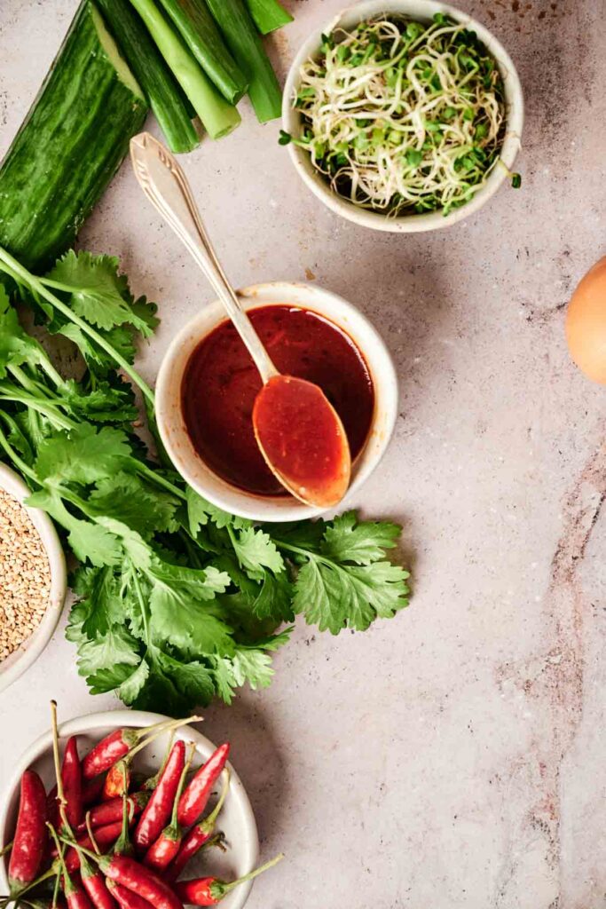 Ingredients on a surface include cilantro, red chili sauce in a bowl, alfalfa sprouts, red chilies, cucumber, spring onions, and sesame seeds.