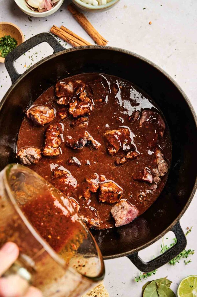 Beef stew simmering in a black pot, with tender chunks of meat and aromatic herbs in a rich brown sauce reminiscent of birria ramen. Onions and other fresh ingredients wait on the side, ready to enhance the hearty dish.