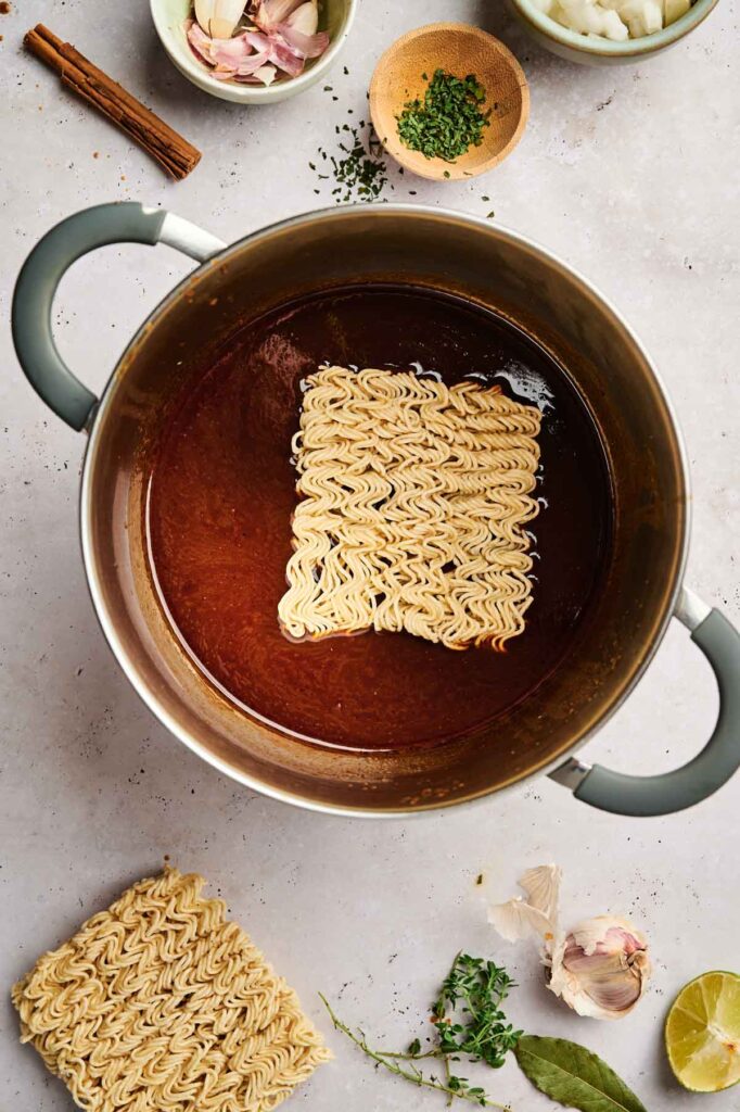 Top view of a pot with birria ramen in a dark broth. Surrounding are ingredients like sliced shallots, herbs, and spices on a light countertop.