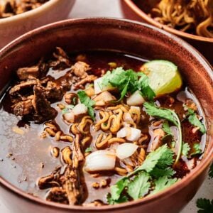 A bowl of savory noodle soup topped with beef, chopped onions, cilantro, and a lime wedge evokes the essence of birria ramen, with another bowl of this flavorful delight in the background.