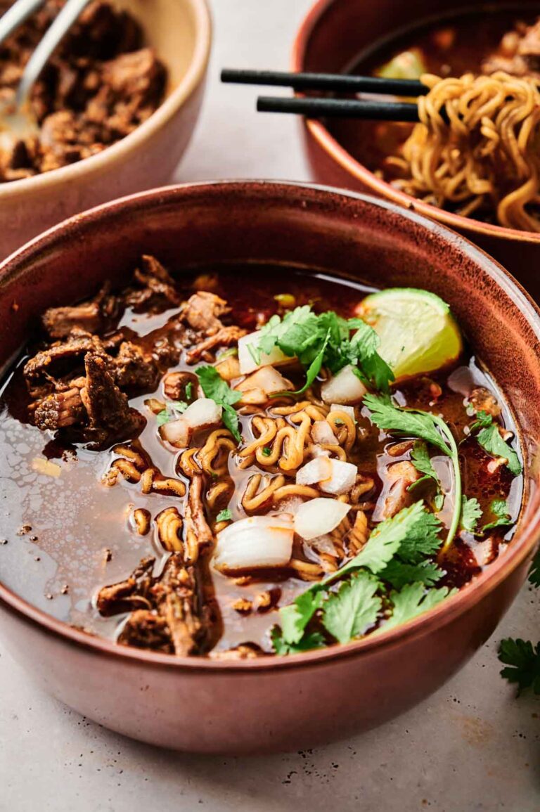 A bowl of savory noodle soup topped with beef, chopped onions, cilantro, and a lime wedge evokes the essence of birria ramen, with another bowl of this flavorful delight in the background.