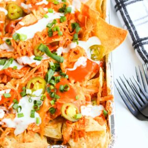 A tray of Loaded Buffalo Chicken Nachos piled high with shredded chicken, melted cheese, jalape&ntilde;os, green onions, hot sauce, and sour cream sits invitingly with a black fork and napkin beside it.