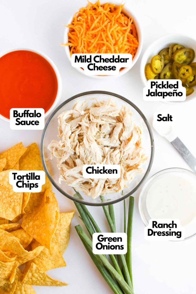 Ingredients laid out for loaded buffalo chicken nachos: tortilla chips, shredded chicken, buffalo sauce, cheddar cheese, pickled jalape&ntilde;os, ranch dressing, salt, and green onions.