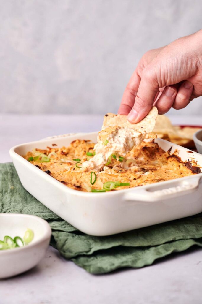 A hand dips a chip into a baked cheese and onion dip in a white dish on a green cloth. Nearby is a small bowl with sliced green onions.