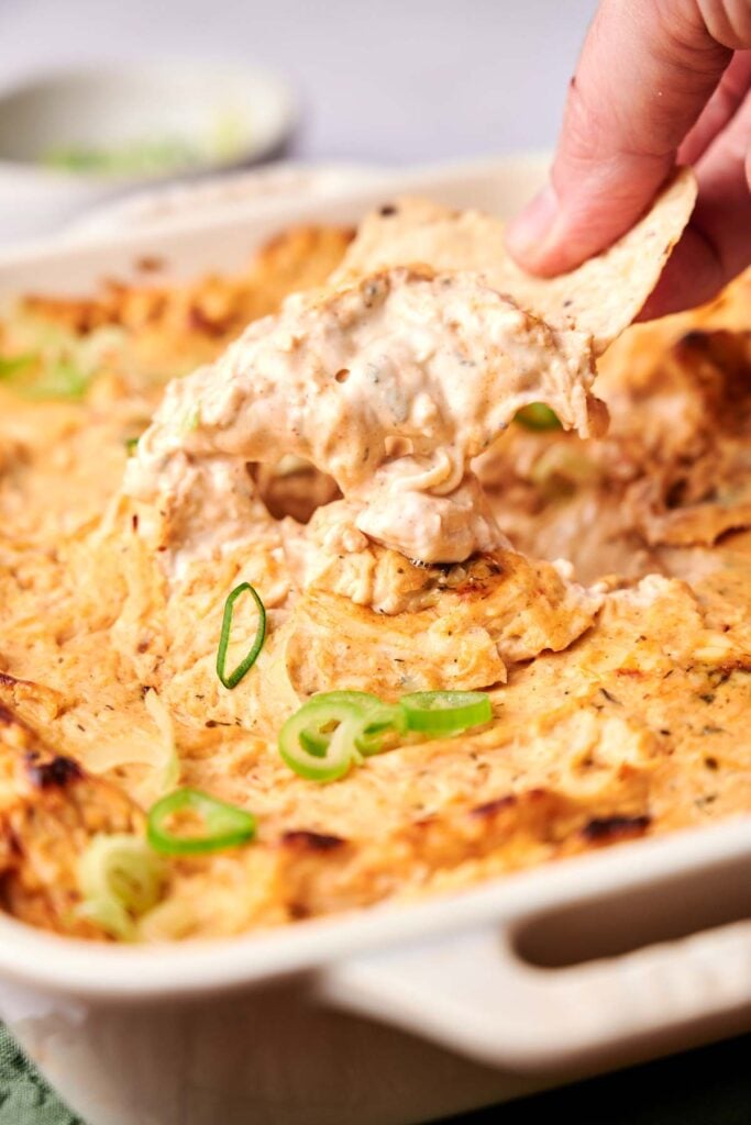 A hand dips a chip into a baked, creamy cheese dip garnished with sliced green onions in a baking dish.