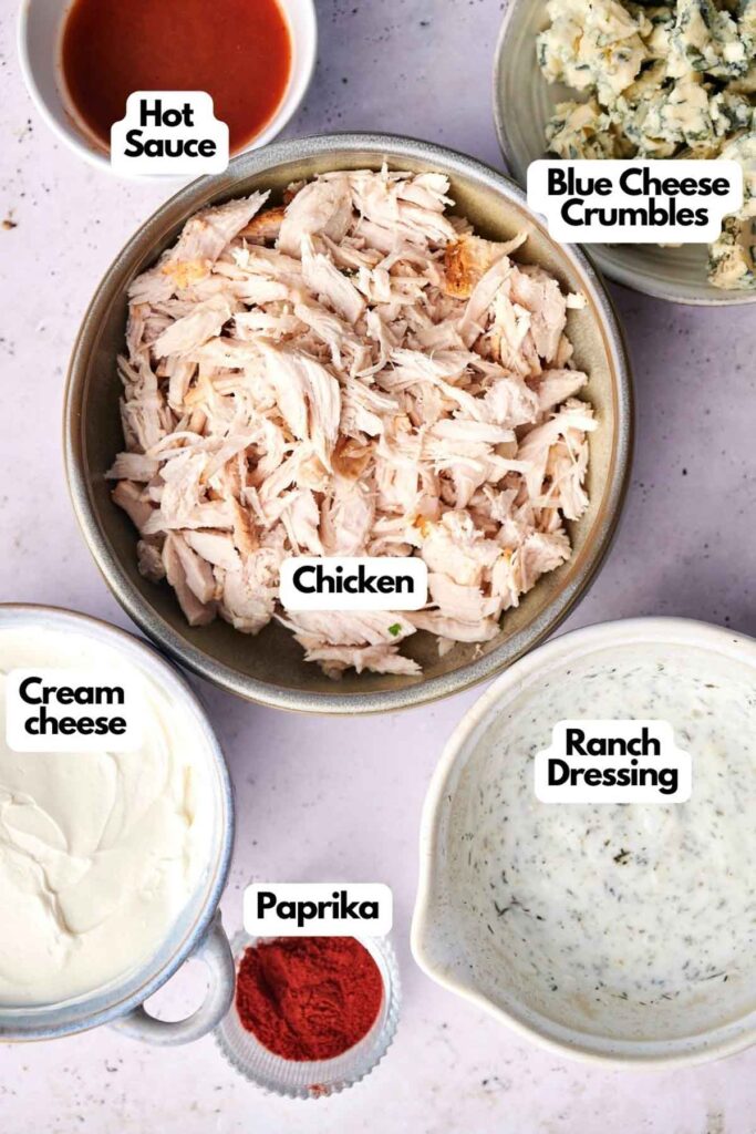 Ingredients for a dish arranged on a surface: shredded chicken in a bowl, hot sauce, blue cheese crumbles, cream cheese, ranch dressing, and paprika.
