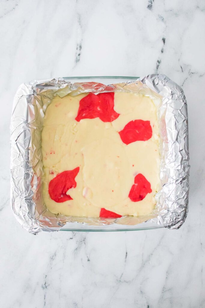 A rectangular baking dish lined with foil contains a candy cane fudge mixture, its red swirls artfully set on the marble countertop.