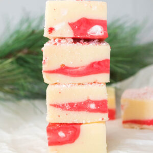 A stack of candy cane fudge swirled with red and white sits on parchment paper, framed by pine branches in the background.