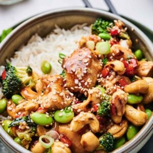 A bowl of Cashew Chicken featuring succulent chicken, broccoli, red peppers, edamame, and sesame seeds sits invitingly, with chopsticks ready in the background.