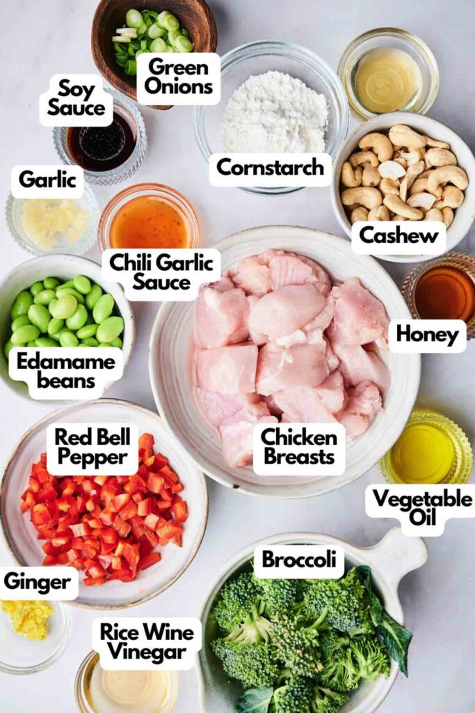 The ingredients for a Cashew Chicken stir-fry are laid out, featuring chicken breasts, broccoli, red bell pepper, and a generous sprinkle of cashews, all ready to be sizzling in soy sauce and cornstarch with various seasonings.