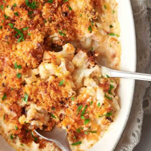 A baked cauliflower gratin with a golden breadcrumb topping, garnished with chopped parsley, displayed in an oval dish with two serving spoons.