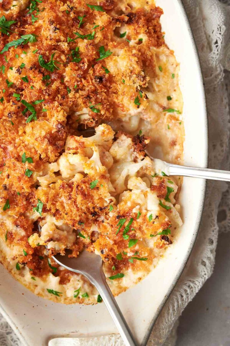 A baked cauliflower gratin with a golden breadcrumb topping, garnished with chopped parsley, displayed in an oval dish with two serving spoons.