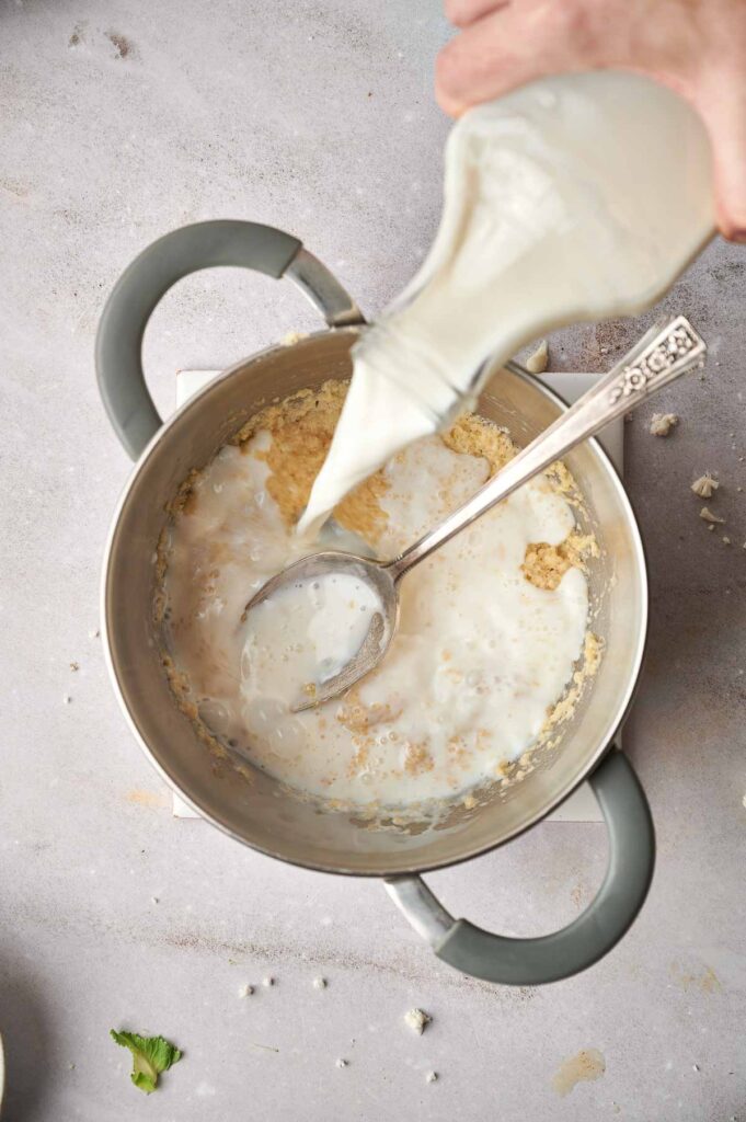 Hand pouring milk from a glass pitcher into a saucepan with cooked oatmeal, next to a spoon, on a kitchen counter.