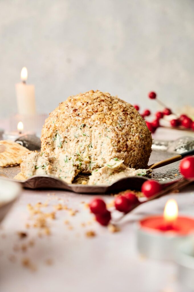 Close-up of a round cheese ball coated in crushed nuts on a wooden board, surrounded by crackers and festive red berries. Candles are lit in the background.