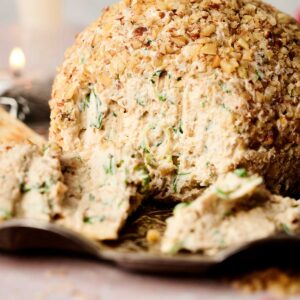 Cheese ball covered in chopped nuts with fresh herbs inside, partially sliced and served on a plate.