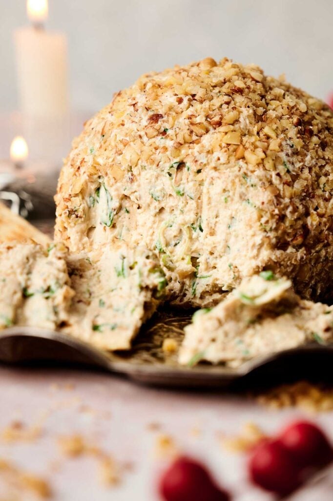 Cheese ball covered in chopped nuts with fresh herbs inside, partially sliced and served on a plate.