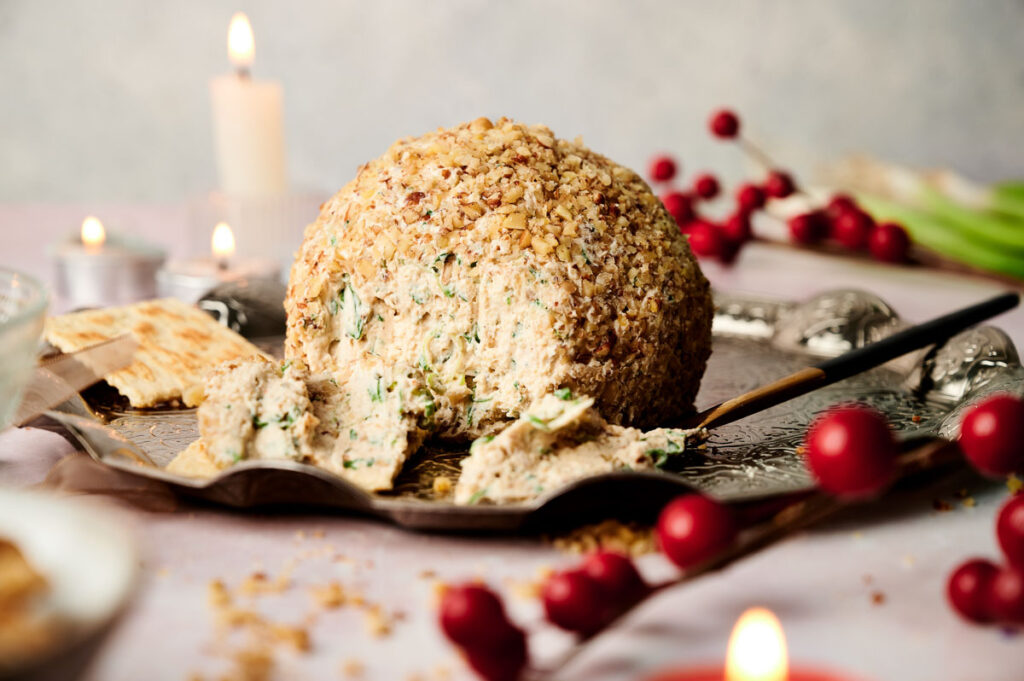 Cheese ball covered in nuts on a tray with serving utensils, surrounded by candles and red berries.