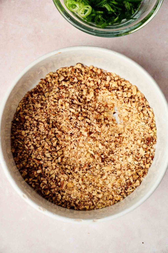A bowl of chopped walnuts is placed on a light surface with a small glass bowl of sliced green vegetables nearby.