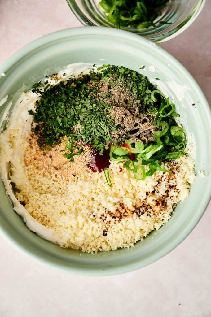 A bowl containing ingredients for a dip, including chopped herbs, grated cheese, mayonnaise, breadcrumbs, sliced green onions, and seasonings.