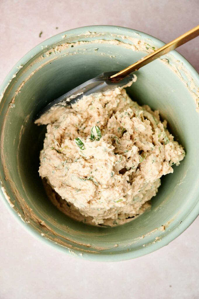 A green bowl contains a creamy mixture with herbs. A gold-handled spatula rests in the bowl.