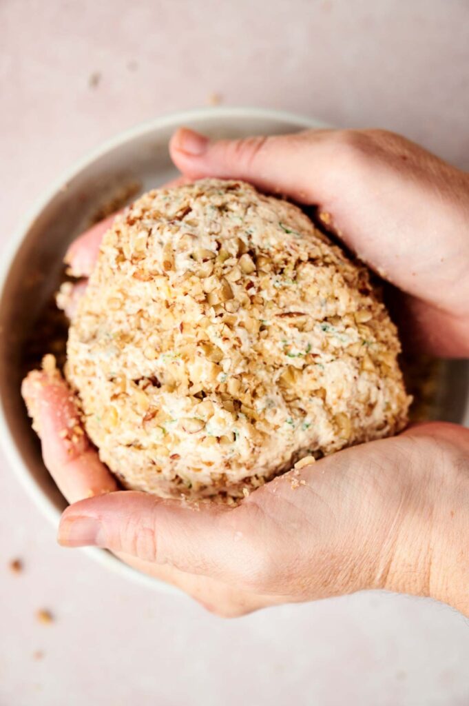 Two hands hold a ball of cheese coated with chopped nuts in a bowl.