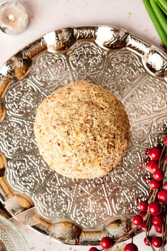 A nut-covered cheese ball on a decorative silver platter, surrounded by red berries and a tea light candle.