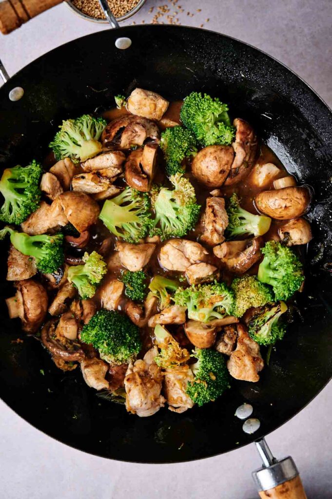 Chicken, broccoli, and mushrooms cooking in a black wok on a light-colored surface.